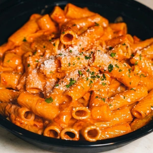 Rigatoni pasta in a creamy orange sauce, topped with grated cheese and parsley, in a black bowl.