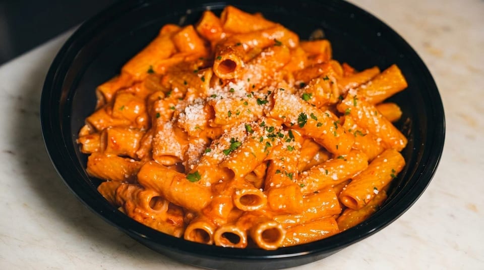Rigatoni pasta in a creamy orange sauce, topped with grated cheese and parsley, in a black bowl.